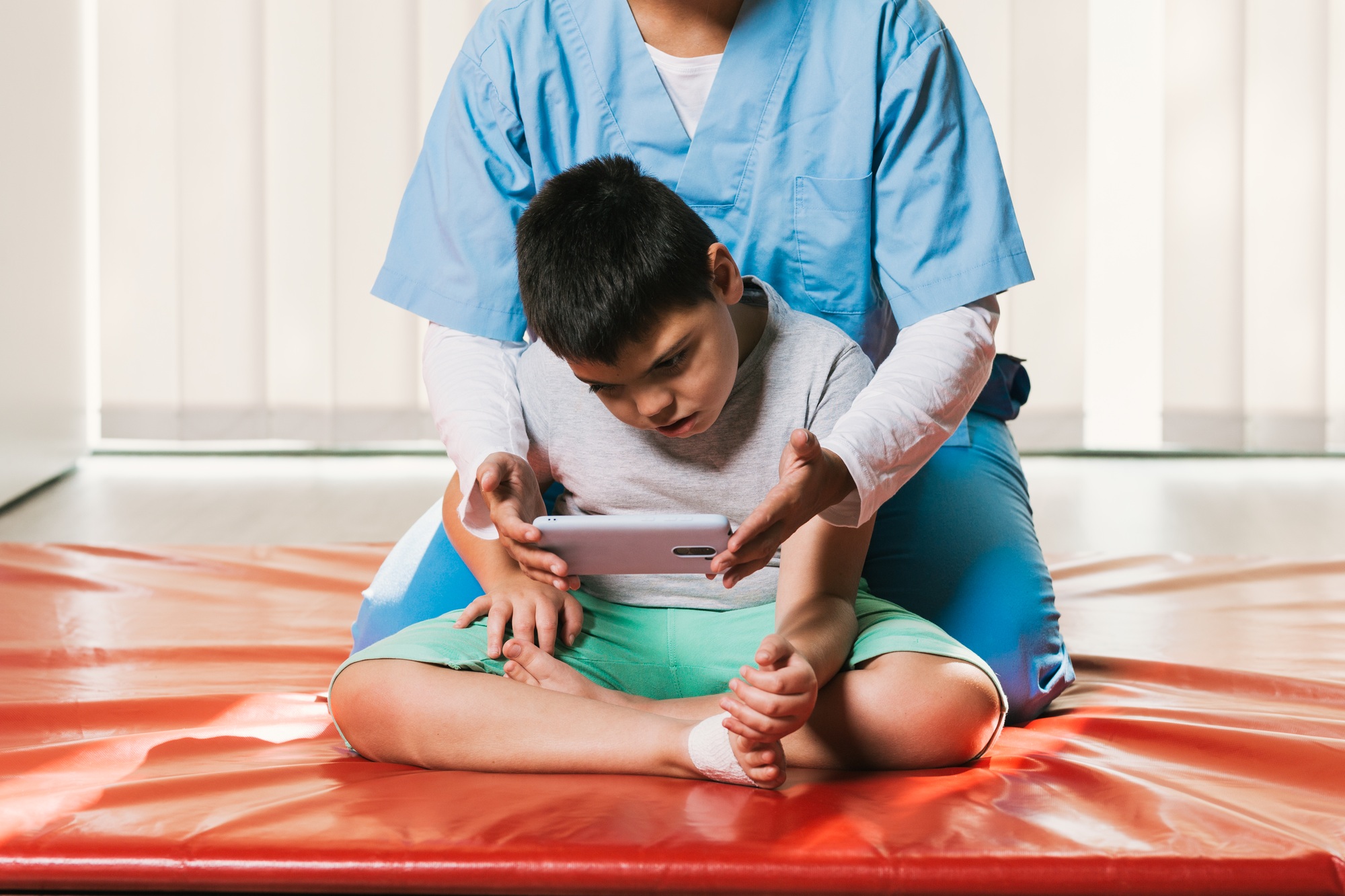 Physiotherapist assisting child with palsy using smartphone for visual tracking and trunk control